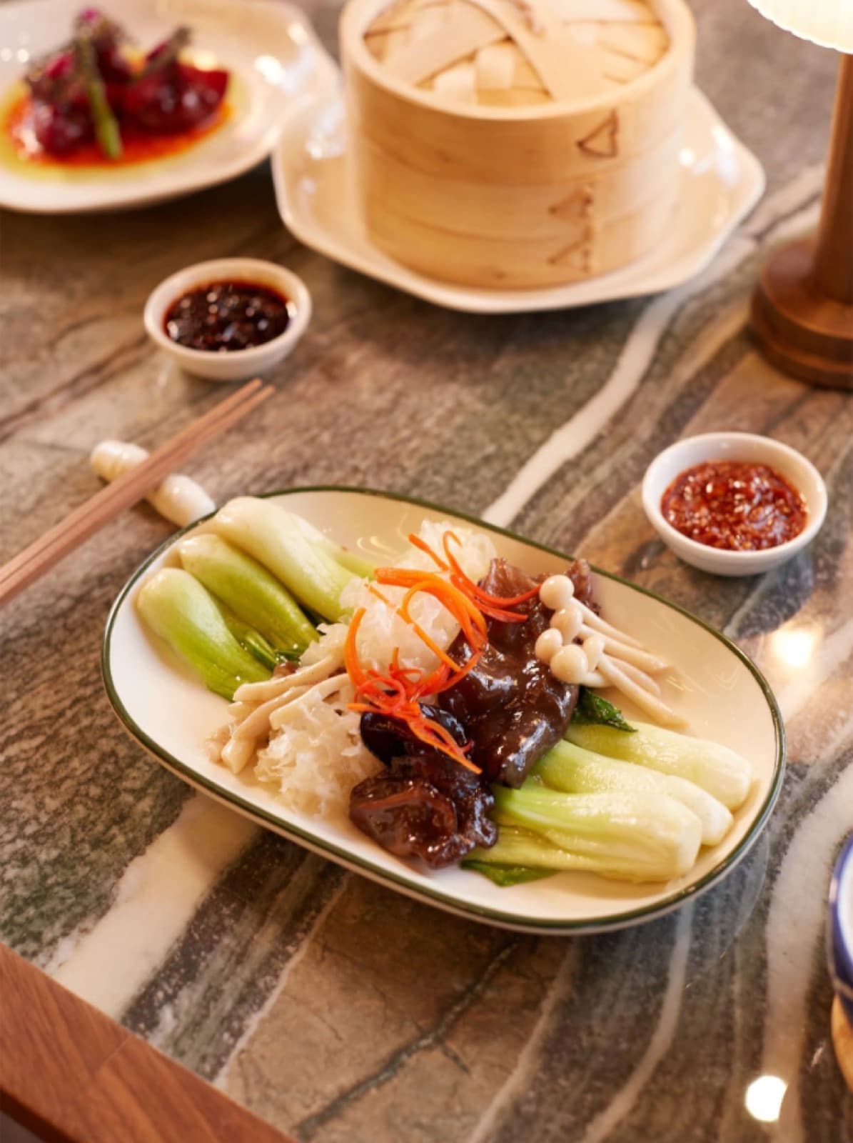 A vibrant plate of steamed bok choy, wood ear mushrooms, and enoki mushrooms garnished with fine carrot slivers, served on a grey marble table alongside a bamboo steamer and small bowls of chilli oil.
