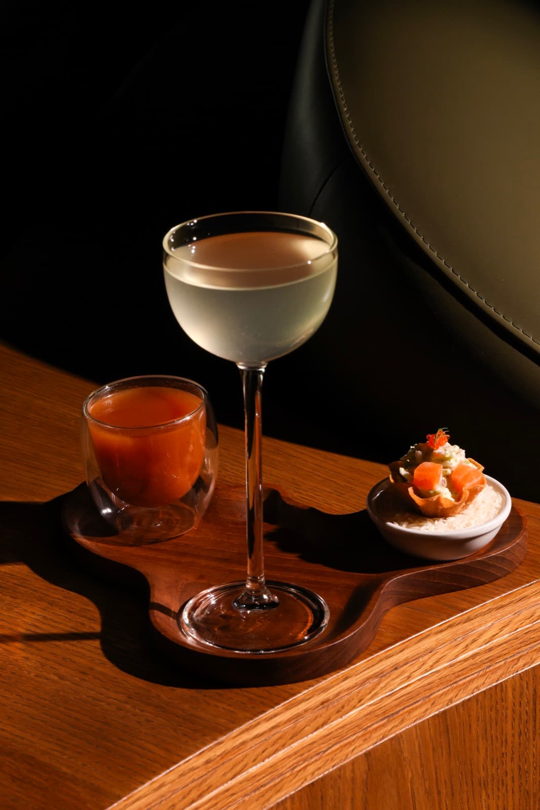 A tall coupe glass of clear cocktail alongside a vibrant michelada chaser and a smoked salmon tartlet, served together on a dark wooden tray at Muro.