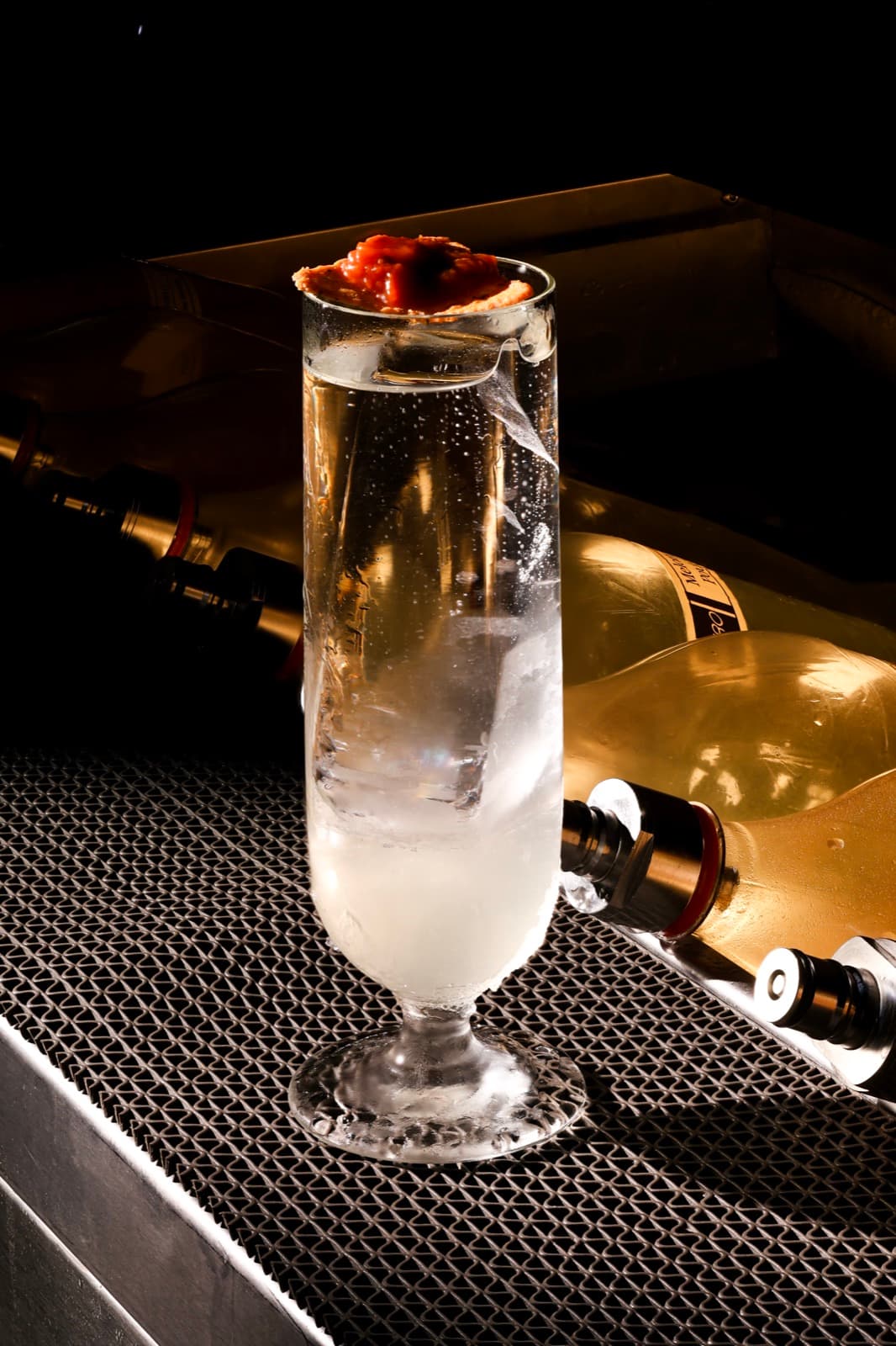 A tall, elegant crystal glass containing the clear El Mariachi cocktail with a large, singular ice spear, topped with a savory red garnish, set against a dark bar background with blurred golden bottles.
