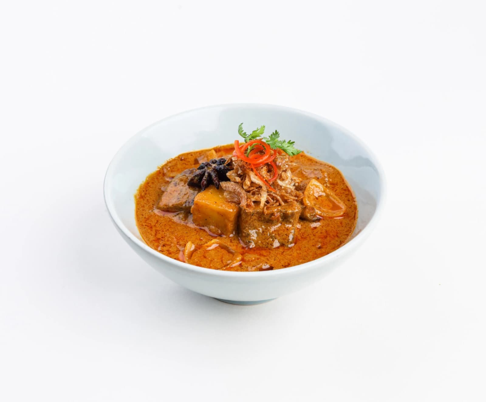 A white bowl containing a rich, orange-hued Thai Massaman Lamb Curry, topped with crispy fried onions, fresh coriander, and a red chilli garnish, isolated on a white background.