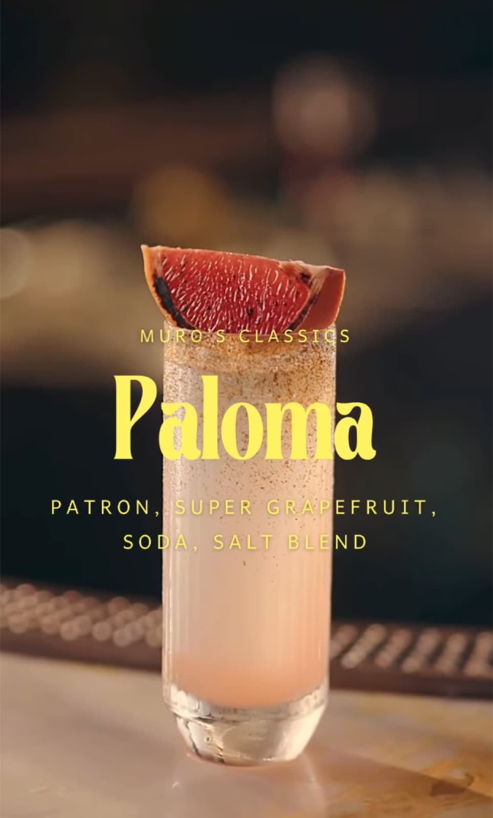 A tall, frosted highball glass filled with a pale pink Paloma cocktail, garnished with a large wedge of charred grapefruit and a salt-dusted rim, set against a warm bar background.