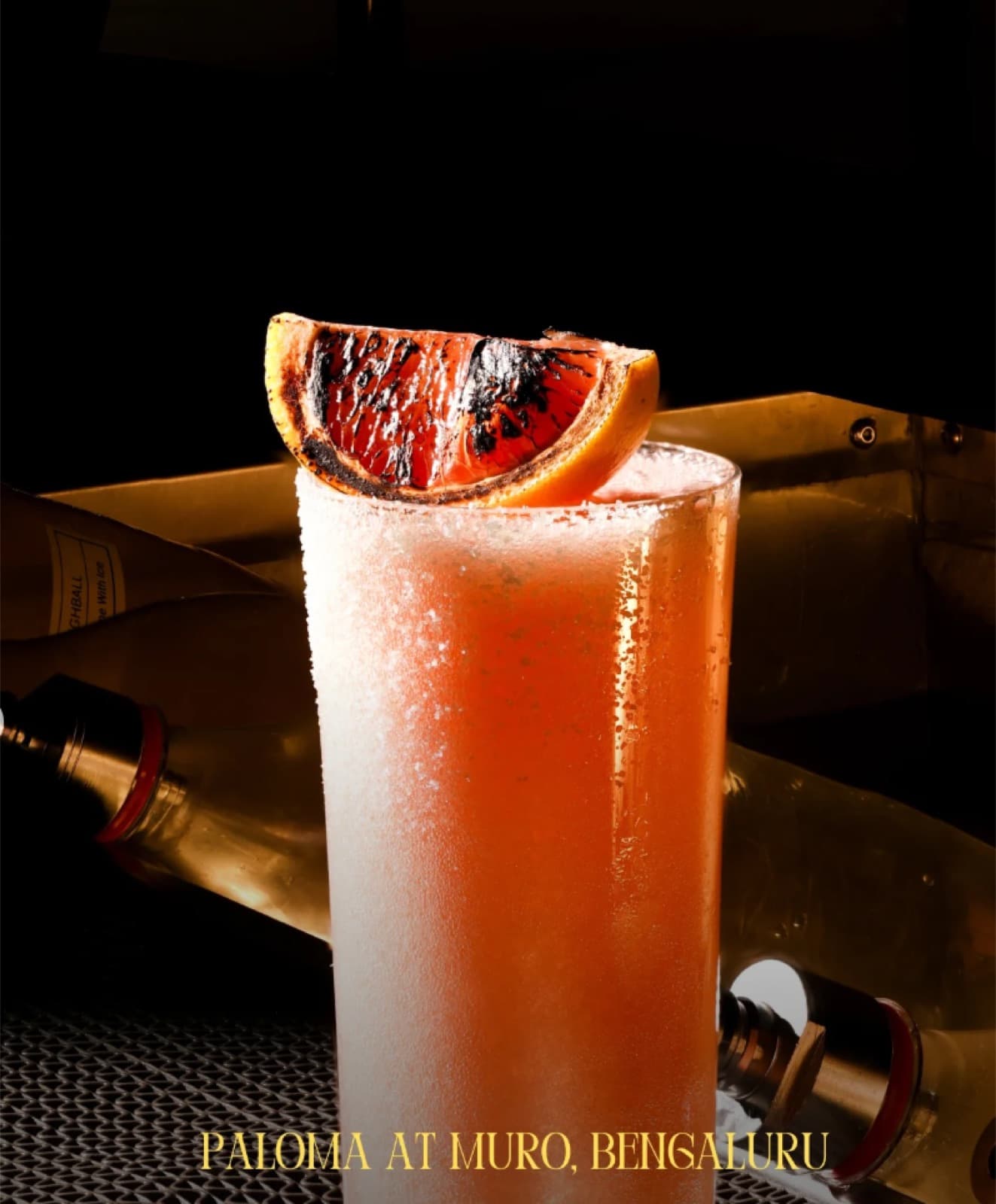 A tall, frosted highball glass of the pink Paloma cocktail at Muro, garnished with a large charred grapefruit slice and served in a moody, upscale bar setting.