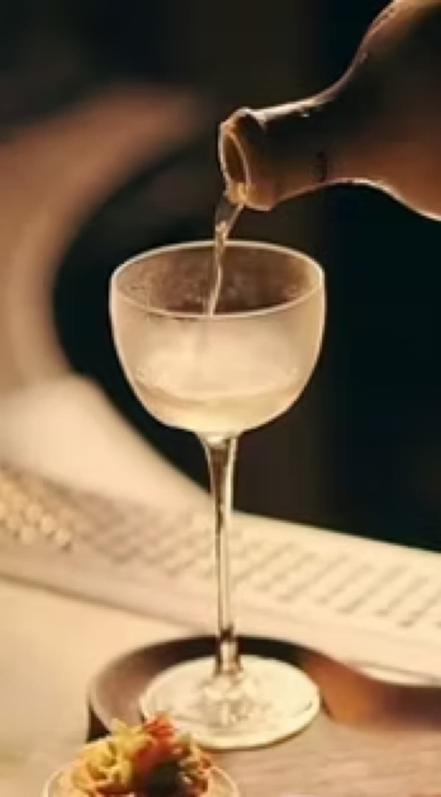 A close-up, warm-toned shot of a clear cocktail being elegantly poured from a dark bottle into a chilled, frosted coupe glass at Muro, Bengaluru.