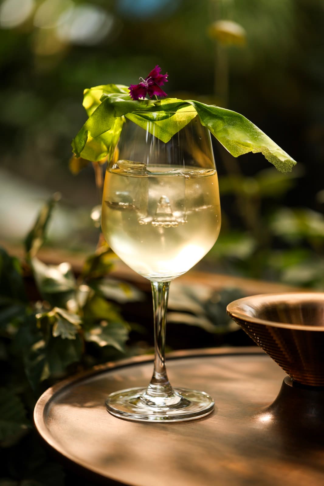A close-up shot of the Shades Off Green cocktail at Muro, served in a wine glass with a large clear ice cube and an elegant green leaf garnish topped with a small purple flower.