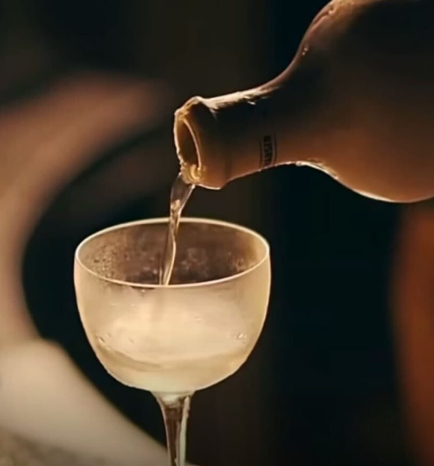 A close-up, warm-toned shot of a clear cocktail being elegantly poured from a dark bottle into a chilled, frosted coupe glass at Muro, Bengaluru.