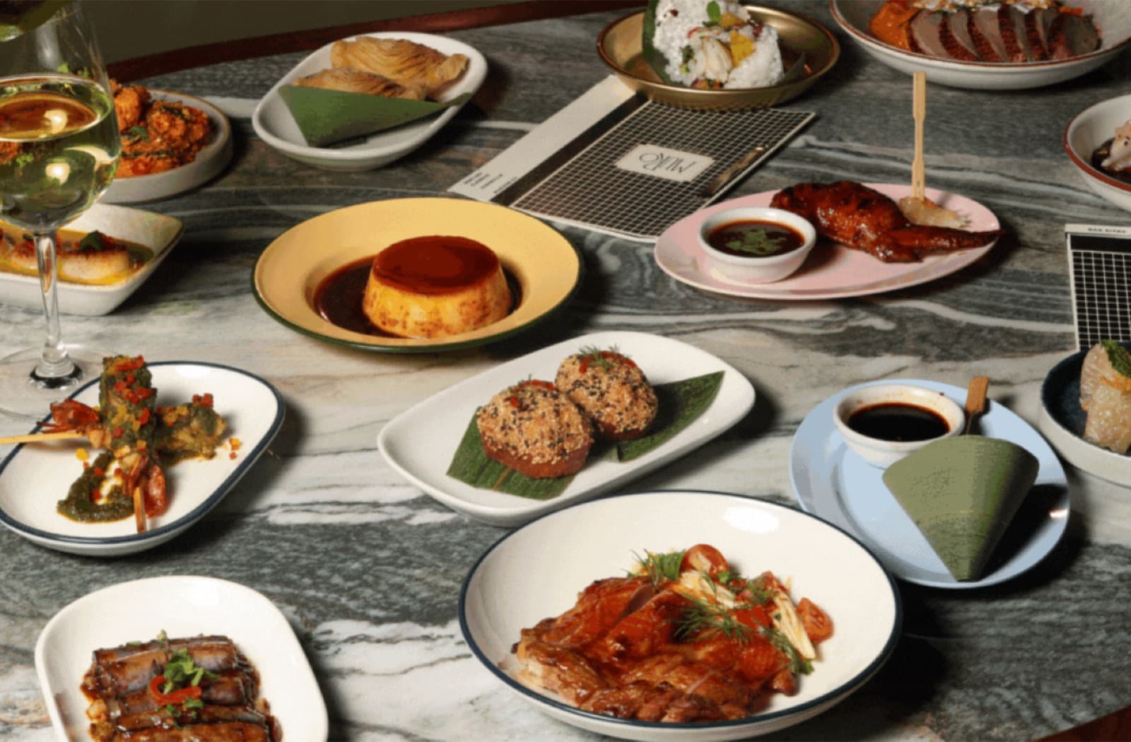 A spread of various Thai and Cantonese-inspired small plates on a grey marble table, including dumplings, grilled meats, and dipping sauces, showcasing the Muro Mezz menu.