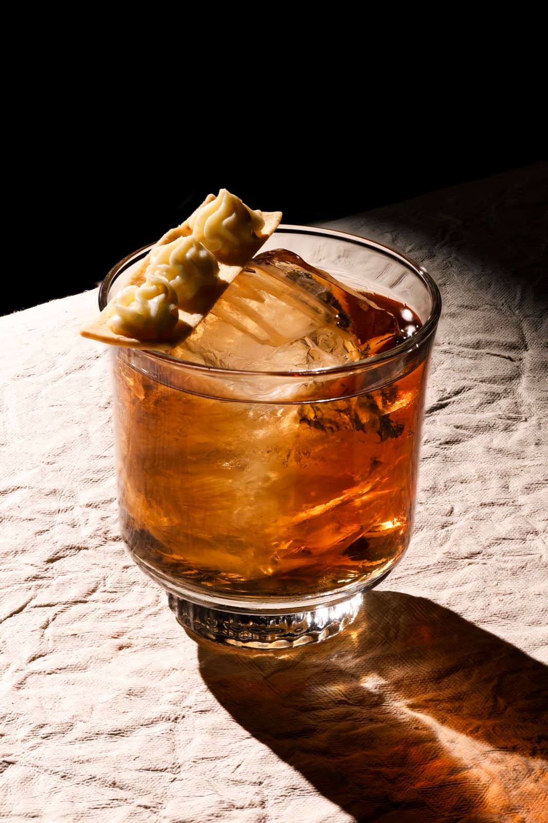 A close-up shot of the Sweet Chin Music cocktail at Muro, showing a crisp lavash garnish topped with three swirls of coconut mousse resting on a large ice cube in a rocks glass.